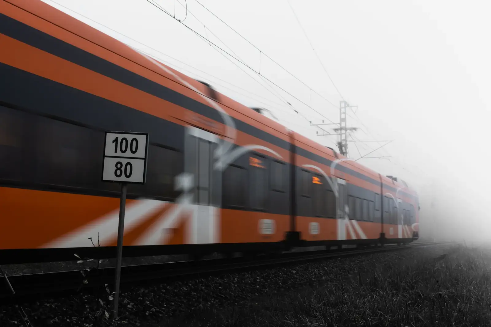 Orange train speeding on railway tracks in dense fog with speed sign visible.